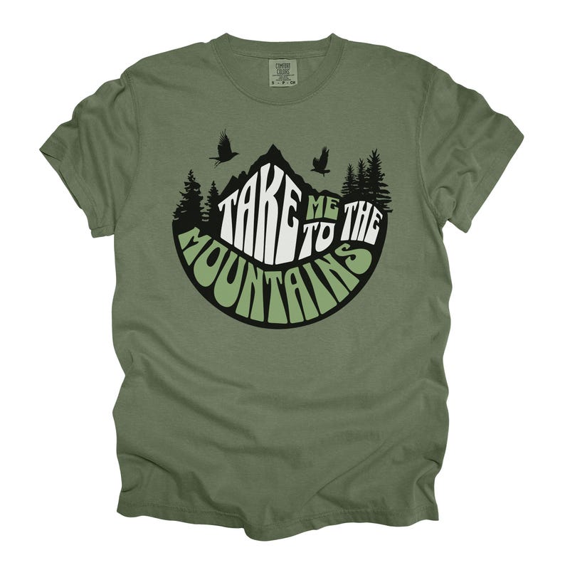 Puede incluir: Camiseta verde oliva con un dise&ntilde;o de monta&ntilde;a. El dise&ntilde;o presenta el texto "TAKE ME TO THE MOUNTAINS" en blanco y verde, con siluetas de &aacute;rboles y p&aacute;jaros. Una camiseta c&oacute;moda e informal.