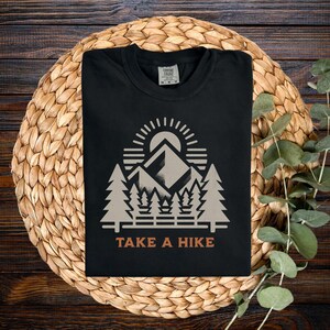May include: Black t-shirt featuring a mountain and forest graphic, a sun, and the text "TAKE A HIKE" in orange. The design includes a mountain range, trees, and a rising sun. The shirt is folded on a woven mat.