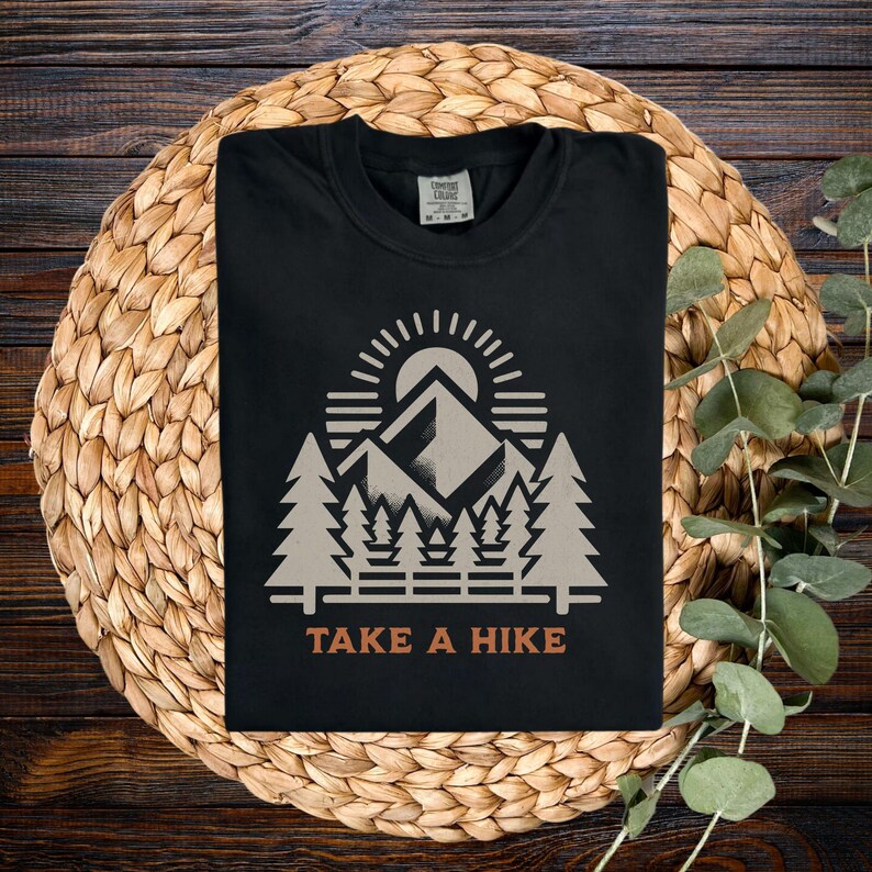 May include: Black t-shirt featuring a mountain and forest graphic, a sun, and the text "TAKE A HIKE" in orange. The design includes a mountain range, trees, and a rising sun. The shirt is folded on a woven mat.