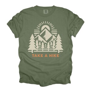 May include: Olive green t-shirt with a white and orange graphic. The graphic depicts a mountain range, sun, and trees, with the text "TAKE A HIKE" underneath. The shirt has a crew neck and short sleeves.