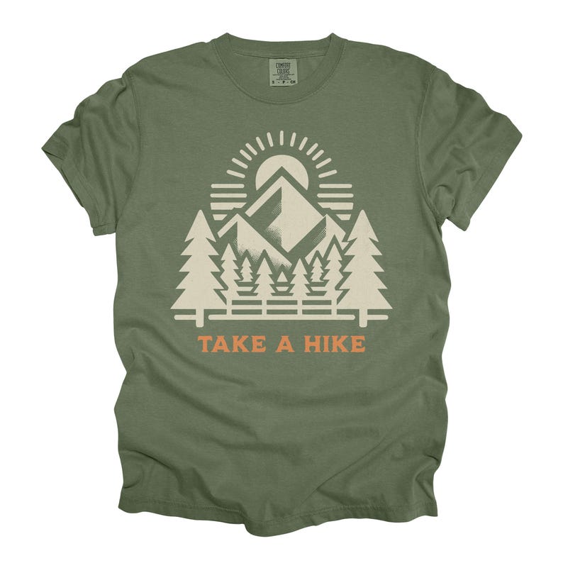 May include: Olive green t-shirt with a white and orange graphic. The graphic depicts a mountain range, sun, and trees, with the text "TAKE A HIKE" underneath. The shirt has a crew neck and short sleeves.