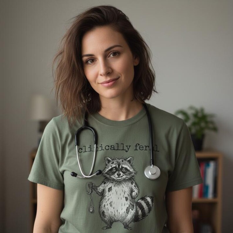 May include: Olive green t-shirt with a raccoon graphic and the text "clinically feral". The raccoon is illustrated in black and white, holding a stethoscope. A black stethoscope is draped around the neck.