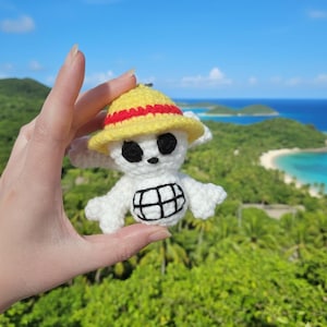 May include: A handmade crochet keychain featuring a white skull with black eyes and a black mouth, wearing a yellow hat with a red band. The keychain is held up against a backdrop of a tropical landscape with green hills and blue ocean.