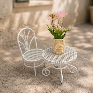 May include: A miniature white metal chair and a matching round table with a mesh surface and scroll legs. A small cylindrical vase wrapped in light tan rope sits on the table, containing a bouquet of artificial pink flowers with green leaves.