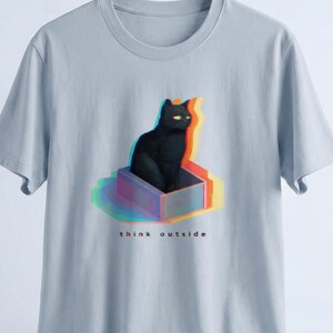 Puede incluir: Camiseta azul claro con un gato negro dentro de una caja con efecto 3D colorido. El texto "think outside" est&aacute; impreso debajo. Cuello redondo y mangas cortas.