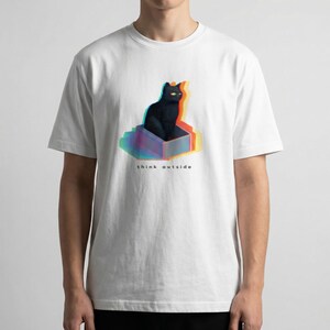 Puede incluir: Camiseta blanca con una ilustraci&oacute;n de un gato negro dentro de una caja con efecto 3D. El gato tiene ojos amarillos y el texto "think outside" est&aacute; debajo. El dise&ntilde;o tiene un efecto colorido y en capas.