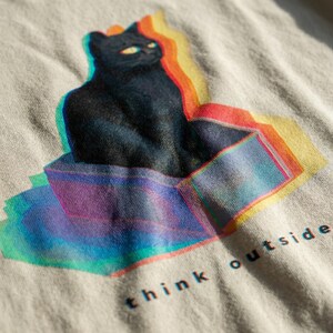 Puede incluir: Camiseta de color claro con una ilustraci&oacute;n de un gato negro con efecto 3D, sentado en una caja. El dise&ntilde;o incluye colores vibrantes como rojo, amarillo, azul y morado. Las palabras "think outside" est&aacute;n impresas debajo.