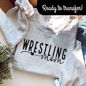 May include: A light gray sweatshirt with the words "WRESTLING mom" in black script. The word "WRESTLING" is in a bold, block font, and "mom" is in a cursive font. The text "Ready to transfer!" is in a black banner at the top.