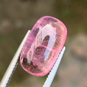Op de afbeelding: Een doorschijnende, roze edelsteen met een rechthoekige vorm, vastgehouden door een zilveren pincet. De edelsteen heeft een gepolijst oppervlak met interne strepen, die het licht reflecteren. De algemene kleur is zachtroze, wat wijst op een toermalijn of een vergelijkbare edelsteen.