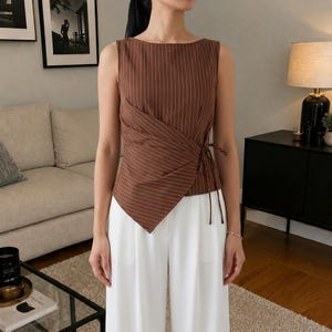 Pode incluir: Uma blusa sem mangas castanha com um padrão de riscas finas brancas, decote barco e uma frente estilo cruzado que se prende com um laço lateral. A peça tem uma bainha assimétrica que se inclina para um lado, criando um efeito sobreposto.