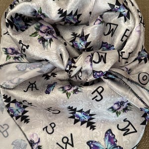 May include: A light gray satin scarf with a paisley pattern, featuring black and purple floral and butterfly designs. The scarf also has black geometric shapes and ranch-style brand symbols. The edges are finished with a black stitched border.