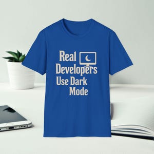 Könnte beinhalten: Königsblaues T-Shirt mit dem weißen Text "Real Developers Use Dark Mode". Das Design zeigt ein Computermonitor-Symbol mit einem Mond. Das T-Shirt wird auf einer weißen Oberfläche präsentiert.