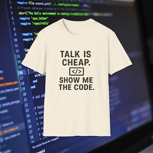 以下が含まれることがあります： 「TALK IS CHEAP. SHOW ME THE CODE.」の黒い文字がプリントされたクリーム色のTシャツ。 コーディング記号が特徴です。 背景にはコード行が表示されたコンピューター画面があります。