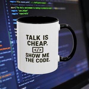 Puede incluir: Taza de café blanca con interior y asa negros. La taza presenta el texto "TALK IS CHEAP. SHOW ME THE CODE." en negro, con un símbolo de código. El fondo muestra código informático.