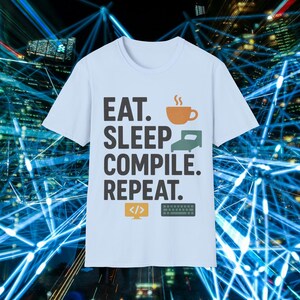 Puede incluir: Camiseta azul claro con la frase "EAT. SLEEP. COMPILE. REPEAT." en negrita. El diseño incluye una taza de café, una cama, una pantalla de ordenador con código y un teclado. La camiseta es de cuello redondo.