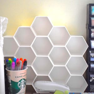 May include: A white, honeycomb-shaped wall shelf unit. The unit is made up of multiple hexagonal compartments. A Starbucks coffee cup with colorful markers and pens sits in front of the shelf. A stapler and other office supplies are also visible.
