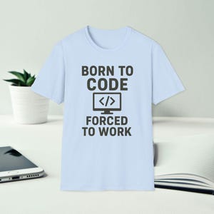 Puede incluir: Camiseta azul claro con el texto "BORN TO CODE" encima de un icono de monitor de ordenador y el texto "FORCED TO WORK" debajo. La camiseta es de cuello redondo y manga corta.