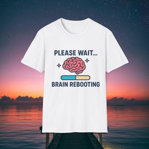 Puede incluir: Camiseta blanca con el texto azul marino "PLEASE WAIT... BRAIN REBOOTING". El diseño incluye un cerebro rosa, una barra de carga azul y amarilla y estrellas.
