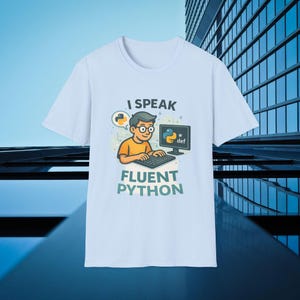 Może przedstawiać: Biała koszulka z grafiką przedstawiającą osobę korzystającą z komputera z napisem "I Speak Fluent Python". Grafika zawiera logo Pythona i słowo "def".