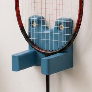 May include: A blue wall-mounted tennis racket holder. The holder is made of plastic and has a U-shaped design to cradle the racket head. The racket is black and red, and the strings are white. The holder is mounted on a white wall.
