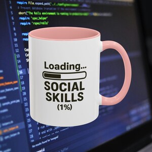 May include: White ceramic mug with a pink interior and handle. The mug features the text "Loading... SOCIAL SKILLS (1%)" in black, with a loading bar graphic. The mug is set against a blurred background.