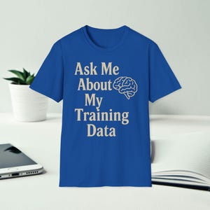 以下が含まれることがあります： 「Ask Me About My Training Data」のテキストがセリフ体で書かれたロイヤルブルーのTシャツ。テキストの右側には白い脳のグラフィックがあります。Tシャツは白い表面に表示されています。