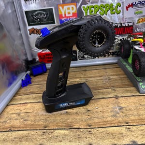 May include: Black FS-GT5 remote control with a blue accent and a black tire attached. The tire has a gold rim and black tread. The remote is on a wooden surface with stickers in the background.