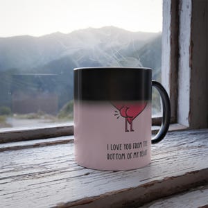 May include: A black and pink color-changing mug with a heart-shaped design and the phrase "I love you from the bottom of my heart." The mug is sitting on a white, weathered wooden surface near a window.