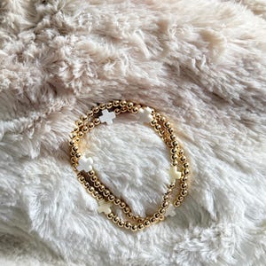 May include: Two gold beaded bracelets with white cross charms. The bracelets are resting on a fluffy, cream-colored surface. The gold beads are small and evenly spaced, and the white crosses are evenly distributed.