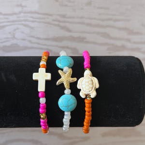 May include: Three beaded bracelets with charms. One bracelet has a white cross charm, another has a gold starfish charm, and the third has a white turtle charm. The bracelets feature pink, orange, and turquoise beads.