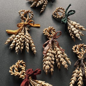 May include: A collection of wheat bundles, each adorned with a ribbon. The bundles have a woven wheat circle at the top and wheat stalks below. Ribbons are in blue, green, brown, burgundy, and gold. The wheat is a natural beige colour.