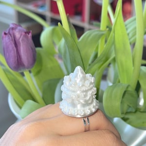 May include: A white, ornate ring with a layered design. The ring is silver and is worn on a finger. The background includes purple tulips and green leaves in a vase.