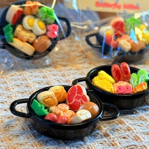 Può includere: Piccole padelle nere piene di cibi colorati e dettagliati. Le padelle contengono uova, carne, verdure e ravioli. Il cibo è disposto in modo da assomigliare a un pasto cucinato. Le padelle hanno piccole maniglie.