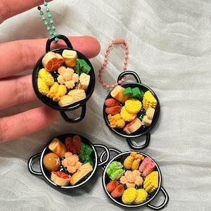 Puede incluir: Llaveros de mini sartenes negras llenas de alimentos coloridos. Cada llavero presenta una pequeña sartén redonda con asas, que contiene varias réplicas de alimentos como carne, verduras y albóndigas. Los llaveros están unidos a cadenas de cuentas.