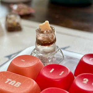 Può includere: Un piccolo bicchiere trasparente con un liquido marrone e un oggetto a forma di orso in cima. Il bicchiere è su un supporto trasparente, con tasti della tastiera rossi e arancioni in primo piano. I tasti hanno testo e simboli bianchi.