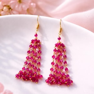 Peut inclure: Boucles d'oreilles pendantes composées de perles rondes rouge rubis. Les perles sont enfilées sur des chaînes dorées, avec des crochets dorés pour oreilles percées. Les boucles d'oreilles sont présentées sur une surface blanche.