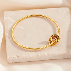 Könnte beinhalten: Ein goldenes Armreif-Armband mit einem Knoten-Anhänger, auf dem die Worte "Forever", "Together" und "Best" eingraviert sind.