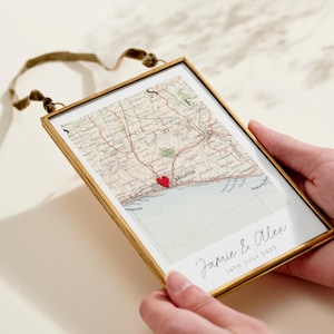 Puede incluir: Impresión enmarcada de un mapa con marco dorado. El mapa muestra una ubicación cerca de Brighton, marcada con un corazón rojo. Los nombres "Jamie & Alex" y la fecha "14th July 2023" están escritos a mano. Una cinta verde está adjunta para colgar.
