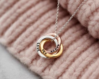 Puede incluir: Un collar de cadena de plata con un colgante de triple anillo. Los anillos están entrelazados y hechos de plata, oro rosa y oro. Los anillos están grabados con las palabras "Mummy", "Love" y "You".
