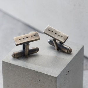 May include: A pair of rectangular silver cufflinks with a series of small, evenly spaced dots etched into the surface. The cufflinks have a tarnished, vintage appearance and are displayed on a gray concrete block.