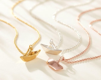 May include: Three necklaces with boat pendants in gold, silver, and rose gold. Each pendant features a small boat design suspended from a delicate chain. The necklaces are displayed on a white surface, with soft lighting.