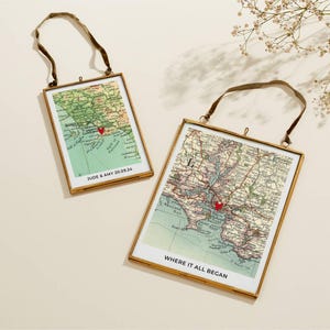 Op de afbeelding: Twee ingelijste vintage kaartprints met goudkleurige metalen frames en bruine fluwelen lintophangers. De kaarten hebben rode hartmarkeringen en de tekst "JUDE & AMY 20.09.24" en "WHERE IT ALL BEGAN". De kaarten hebben een groen, blauw en bruin kleurenschema.