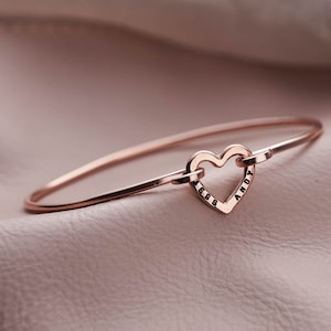 May include: A rose gold bangle bracelet with a heart-shaped charm. The charm has the word "ANDY" and the numbers "1688" engraved on it.