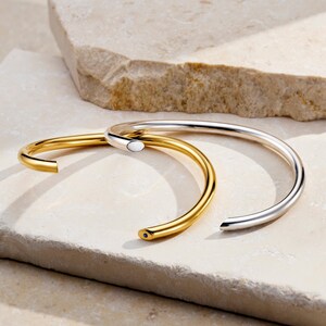 May include: Two open-ended bangles, one gold-toned and one silver-toned, rest on a light-colored stone surface. The gold bangle has a small, dark accent at its end. The silver bangle has a smooth, polished finish, reflecting light.