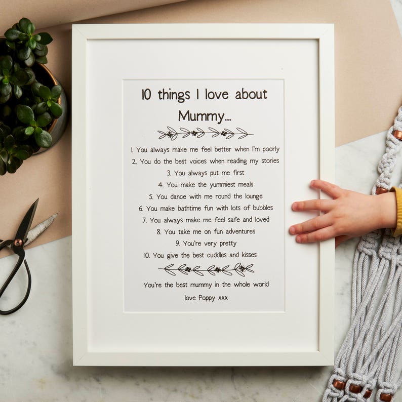 K&ouml;nnte beinhalten: Ein wei&szlig;er gerahmter Druck mit einer Schwarzwei&szlig;illustration einer Kinderhand, die einen Stift h&auml;lt. Der Druck zeigt ein Gedicht mit dem Titel "10 Dinge, die ich an Mama liebe..." mit einer Liste von Gr&uuml;nden, warum ein Kind seine Mutter liebt. Das Gedicht endet mit "Liebe Poppy xxx".