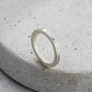 Op de afbeelding: Een zilveren ring met een getextureerd oppervlak en kleine, donkere markeringen. De ring staat tegen een lichtgrijze, getextureerde achtergrond. Het ontwerp van de ring is eenvoudig en modern.
