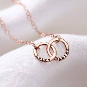 May include: A rose gold necklace with two interlocking circles, each engraved with a name: "Arley" and "Daniel".