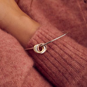 以下が含まれることがあります： 金と銀が絡み合ったリングチャームが付いたシルバーのバングルブレスレット。チャームには「Elle」、「Flex」、「Bair」という名前が刻まれています。