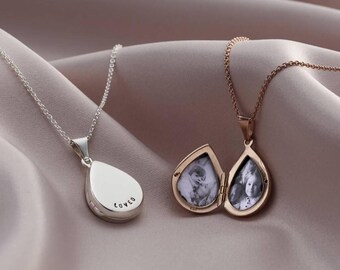 May include: Two teardrop-shaped locket necklaces. One is silver with the word "LOVED" engraved on the side. The other is gold-toned and open, revealing two black and white photographs. Both necklaces hang on delicate chains.