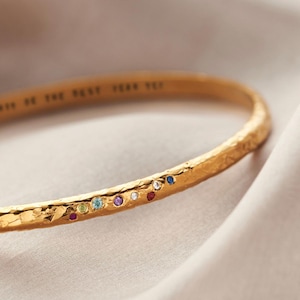 May include: A gold bangle bracelet with a textured finish. The bracelet features a line of small, multi-coloured gemstones. The inscription "...BE THE BEST YEAR YET" is engraved on the bracelet. The bracelet is presented on a soft, neutral-toned fabric.